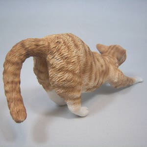 Puede incluir: Una figurita de cer&aacute;mica de un gato jengibre estir&aacute;ndose con la cola enroscada alrededor de su cuerpo.