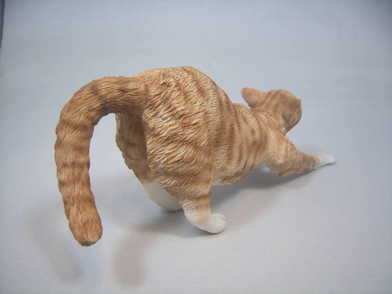 Puede incluir: Una figurita de cer&aacute;mica de un gato jengibre estir&aacute;ndose con la cola enroscada alrededor de su cuerpo.