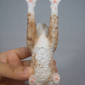 Puede incluir: Una figurita de gato blanco y naranja con almohadillas rosas, colgando boca abajo con las patas en el aire.