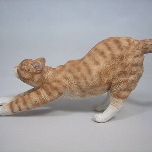 Puede incluir: Una figurita de cer&aacute;mica de un gato jengibre estir&aacute;ndose con la espalda arqueada y la cola enroscada. El gato est&aacute; pintado con detalles realistas, incluyendo rayas y un pecho blanco.