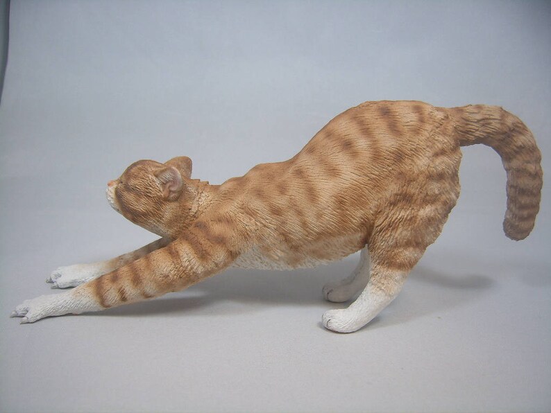 Puede incluir: Una figurita de cer&aacute;mica de un gato jengibre estir&aacute;ndose con la espalda arqueada y la cola enroscada. El gato est&aacute; pintado con detalles realistas, incluyendo rayas y un pecho blanco.