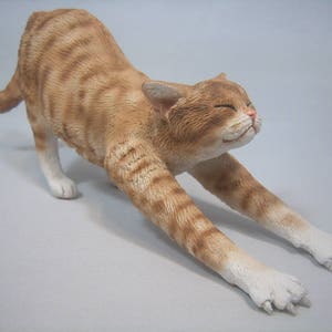 Puede incluir: Una figurita de cer&aacute;mica de un gato jengibre estir&aacute;ndose con las patas extendidas y la cabeza echada hacia atr&aacute;s. El gato tiene patas blancas y un pecho blanco.