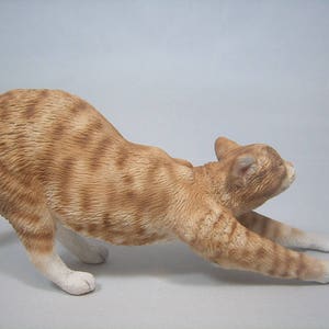 Puede incluir: Una figurita de cer&aacute;mica de un gato jengibre estir&aacute;ndose con las patas extendidas. El gato tiene un pecho y patas blancas.