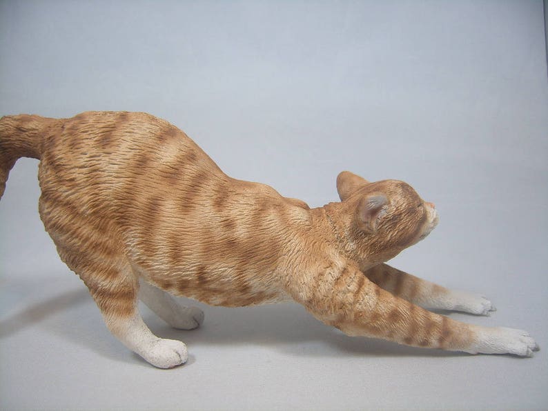 Puede incluir: Una figurita de cer&aacute;mica de un gato jengibre estir&aacute;ndose con las patas extendidas. El gato tiene un pecho y patas blancas.