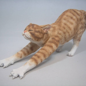 Puede incluir: Una figurita de cer&aacute;mica de un gato jengibre estir&aacute;ndose con patas blancas y un cuerpo rayado marr&oacute;n y blanco.