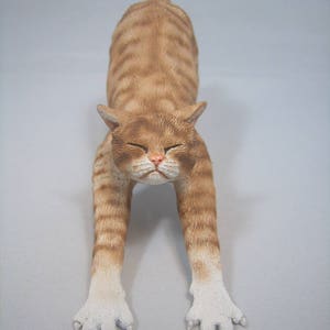 Puede incluir: Una figurita de cer&aacute;mica de un gato jengibre estir&aacute;ndose con las patas extendidas hacia adelante y la cabeza inclinada hacia abajo. El gato tiene patas blancas y un pecho blanco.