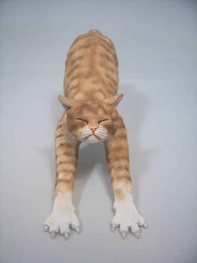 Puede incluir: Una figurita de cer&aacute;mica de un gato jengibre estir&aacute;ndose con las patas extendidas hacia adelante y la cabeza inclinada hacia abajo. El gato tiene patas blancas y un pecho blanco.