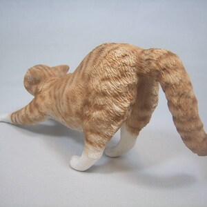 Puede incluir: Una figurita de cer&aacute;mica de un gato jengibre estir&aacute;ndose con la cola enroscada alrededor de su cuerpo.