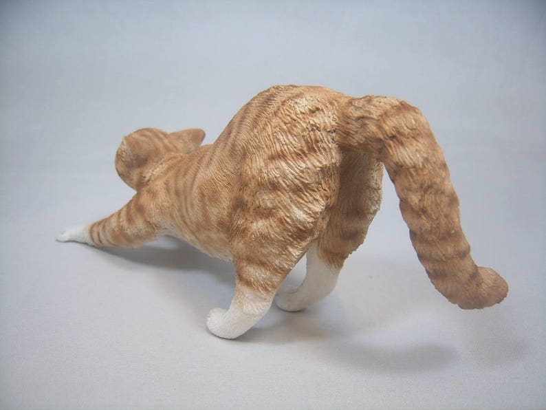 Puede incluir: Una figurita de cer&aacute;mica de un gato jengibre estir&aacute;ndose con la cola enroscada alrededor de su cuerpo.