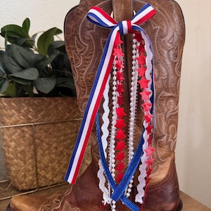May include: Brown leather cowboy boot with decorative red, white, and blue ribbons and star accents. The boot has a pointed toe and intricate stitching. A patriotic-themed accessory for a Western-style boot.