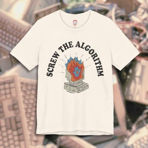 May include: Cream-colored t-shirt featuring the phrase "SCREW THE ALGORITHM" in black, arched above a retro computer graphic with flames. The graphic is in shades of red, orange, blue, and gray. The shirt has a small logo on the upper back.