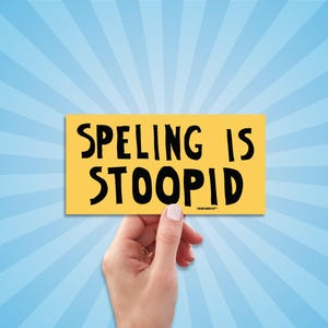 May include: A yellow rectangle with the words "SPELING IS STOOPID" in black, held by a hand against a blue and white radial background. The text is in a bold, sans-serif font.