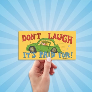 May include: A yellow rectangle with the words "DON'T LAUGH IT'S PAID FOR!" in red and blue. A green cartoon car with a driver is depicted. The image is held by a hand against a blue and white striped background.