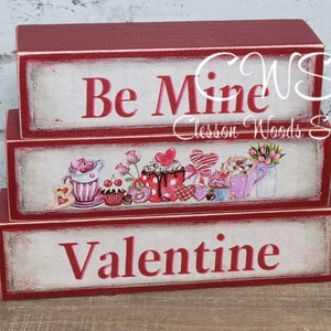 May include: Three stacked wooden blocks with a Valentine's Day theme. The top block is red and says "Be Mine". The middle block has a white background with a colorful illustration of teacups, cupcakes, and hearts. The bottom block is red and says "Valentine".