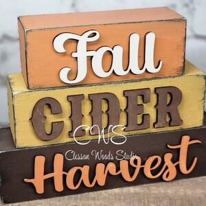 May include: Stacked wooden blocks with the words "Fall Cider Harvest" in a rustic style. The blocks are painted in shades of orange, yellow, and brown.