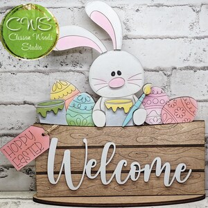 May include: A wooden sign featuring a white bunny holding a paintbrush, surrounded by colorful painted eggs. The sign has a wood grain texture with the word Welcome in white script and a pink tag that reads Hoppy Easter.