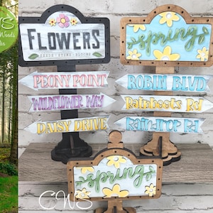 May include: Wooden signs with a spring and flower theme. The signs have painted arrows with names like "Peony Point", "Wildflower Way", "Daisy Drive", "Robin Blvd", "Rainboots Rd", and "Raindrop Rd".