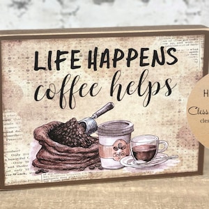May include: A brown wooden sign with the text "Life Happens coffee helps" in black lettering. The sign features a vintage-style illustration of a bag of coffee beans, a scoop, a cup of coffee, and a coffee mug. A brown paper tag with the text "Handmade by Clesson Woods Studio clessonwoodsstudio.com" is attached to the sign.