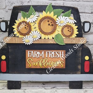 May include: A black wooden truck cutout featuring a bed filled with yellow sunflowers and white daisies. A rectangular sign on the tailgate displays the text FARM FRESH Sunflowers in white and yellow lettering. The truck has a gray bumper.