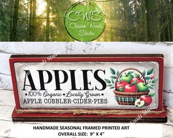 May include: A rectangular, framed wooden sign with a red border features the word "APPLES" in large black letters. Below, text reads "100% Organic Locally Grown" and "APPLE COBBLER CIDER PIES." A basket of apples is depicted. Overall size: 9" x 4".
