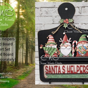 May include: A black wooden bread board with a shelf and three painted gnomes. The gnomes are wearing red, green, and blue hats and holding a Christmas tree, candy cane, and a red and white ball. The board has the text "Santa's Helpers" in red and white letters and the text "Elf Village - Reindeer Games - Workshop" in black letters.