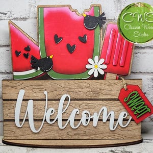 May include: A wooden tabletop sign featuring a watermelon slice, a red popsicle, and two black birds with hearts. The base is a light brown wood-grain plaque with the white text Welcome and a red tag hanging from the side with the green text Sweet.