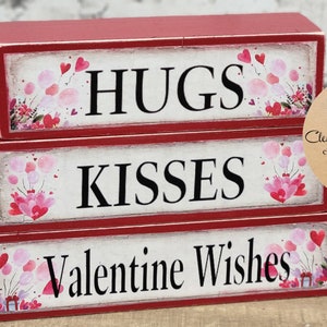 May include: Three stacked wooden blocks with a red background and white lettering. The top block says "HUGS", the middle block says "KISSES", and the bottom block says "Valentine Wishes". Each block has a pink and white watercolor design with hearts and flowers.
