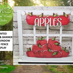 May include: A white wooden window frame display featuring a hanging banner with the word APPLES in white lettering. The display includes a stack of red painted wooden apples with green leaves attached to the frame and base.