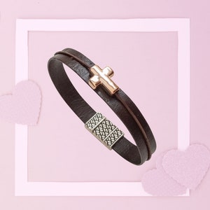 May include: A dark brown leather bracelet with a gold-coloured cross charm. The bracelet has a silver-coloured magnetic clasp. The background is a light pink with white heart-shaped decorations.