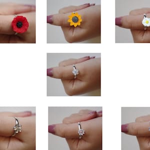 May include: A selection of floral rings with silver bands and colourful flower designs. The rings showcase a red poppy, yellow sunflower, white daisy, and other blossoms. The rings are displayed on a finger. The flowers are in various colours.