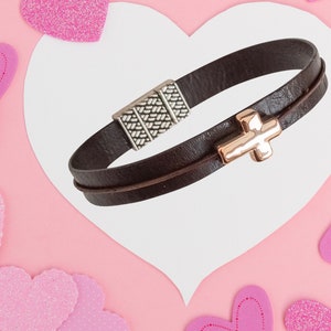 May include: A dark brown leather bracelet with a silver clasp and a gold cross charm. The bracelet is displayed on a white heart-shaped card against a pink background with pink heart decorations.
