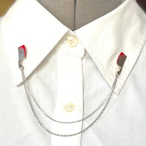 May include: A white collared shirt with two silver chains attached to small red and silver knife-shaped collar clips.