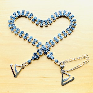 May include: A light blue rhinestone bra strap with two silver metal clasps. The strap is made of two rows of rhinestones connected by silver metal.