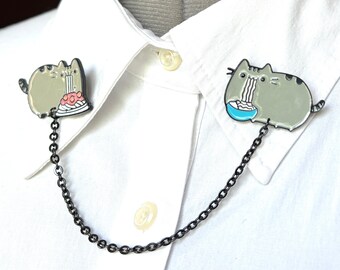 May include: A black chain collar clip with two enamel pins featuring a cartoon cat eating a cake and a cartoon cat eating noodles.