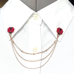 May include: A gold chain collar pin with two red and black ladybug charms. The chain is made of three strands of different sizes.