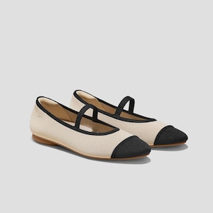 May include: A pair of ballet flats in a neutral color with black accents. The shoes have a rounded toe, a strap across the top, and a black trim. The soles are a light brown color.