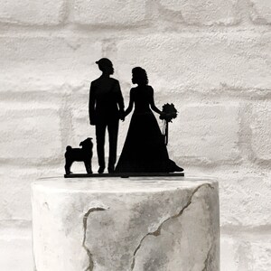 May include: Black silhouette wedding cake topper depicting a couple, a bride with a bouquet, and a small dog. The topper is on a cake with a marbled grey and white design.