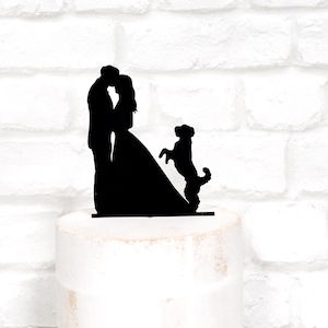 May include: Black silhouette cake topper featuring a kissing couple and a dog standing on its hind legs. The bride is wearing a long dress. The topper is placed on a white cake, with a white brick background.