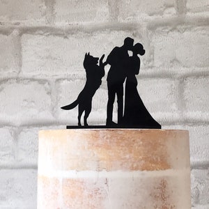 May include: Black silhouette cake topper featuring a couple kissing and a dog jumping up. The couple is standing on a wooden base.