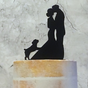 May include: Black silhouette cake topper of a couple kissing with a dog standing in front of them. The couple is standing on a cake with a brick pattern.