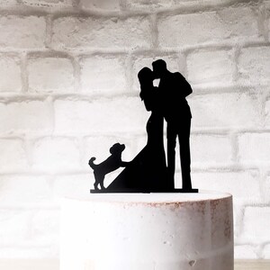 May include: Black silhouette cake topper of a couple kissing with a dog standing next to them. The couple is facing each other and the dog is looking at the couple.