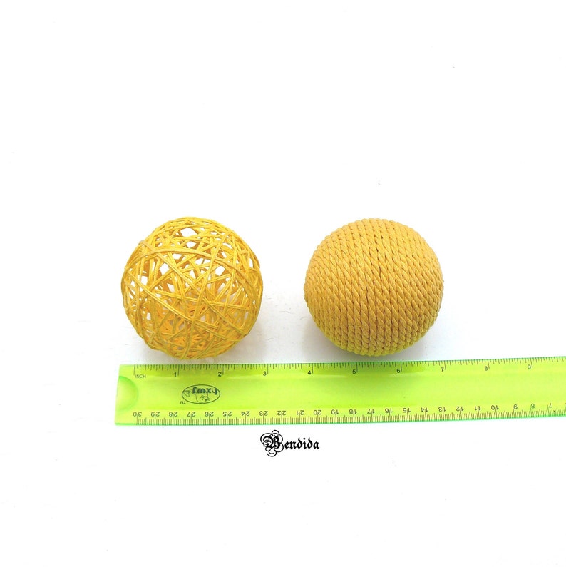 May include: Two yellow woven spheres, one with a more open weave and the other with a tighter weave, are shown on a white surface with a green ruler for scale.