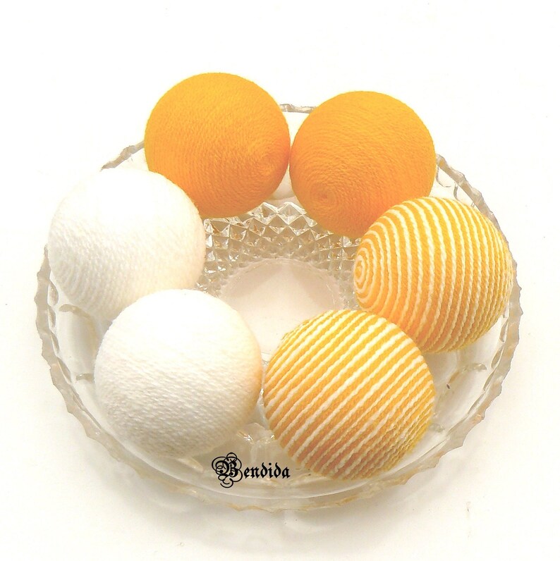 May include: A glass bowl filled with six cotton balls. The cotton balls are in two colors: white and yellow. The yellow cotton balls have a striped pattern.