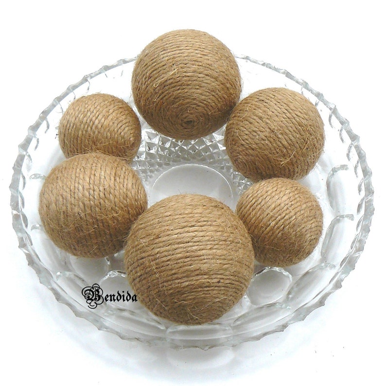 May include: Six brown jute twine balls in a clear glass bowl with a diamond pattern. The balls are made of natural twine and are a popular decorative element for home decor.