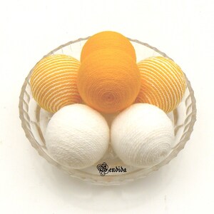 May include: A glass bowl filled with six yarn balls. There are two white yarn balls, two yellow yarn balls, and two yellow and white striped yarn balls.