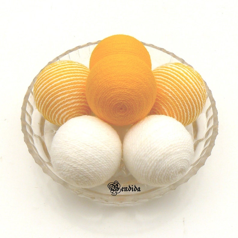 May include: A glass bowl filled with six yarn balls. There are two white yarn balls, two yellow yarn balls, and two yellow and white striped yarn balls.