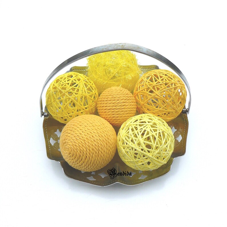 May include: A silver wire basket with a handle holds six yellow yarn balls. The balls are different sizes and textures, some are smooth and some are woven.