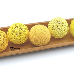 May include: Four yellow woven balls and one yellow fluffy ball in a wooden tray. The balls are made of natural fibres and have a rustic look.