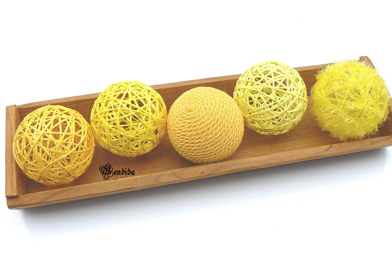 May include: Four yellow woven balls and one yellow fluffy ball in a wooden tray. The balls are made of natural fibres and have a rustic look.