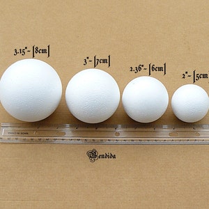 May include: Four white styrofoam balls of different sizes, measured in inches and centimeters, on a brown surface with a ruler.